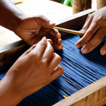 **Prompt 1: "The Art of Natural Dyes and Fibers"**
    A close-up, warm-toned photograph of a skilled Thai artisan's hands, deeply engrossed in crafting. Their hands are working with naturally dyed textiles, showcasing earthy tones of indigo, madder, and turmeric, or intricately weaving delicate strands of water hyacinth or banana fiber. The background reveals a traditional Thai workshop setting, bathed in soft, natural light, with elements like stacks of sustainable materials (bamboo, coconut shells, plant-based dyes) and lush tropical foliage visible through an open window. Emphasize the texture of the materials and the artisan's focused, respectful interaction with nature, embodying ancestral wisdom and ecological consciousness. Detailed, authentic, soulful, cinematic lighting.